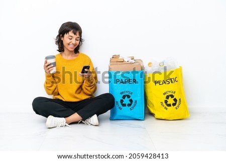 Young girl holding a bag full of plastic and paper sitting on the floor isolated on white background holding coffee to take away and a mobile