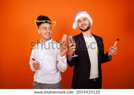 New year, party, homosexuality and people concept - Happy two gay international friends with a Santa hat and star glasses with party horns and champagne glasses on an orange background with copy space