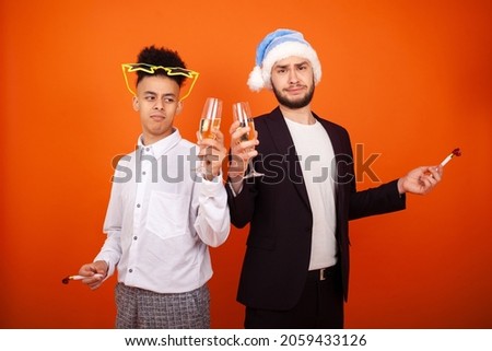 New year, celebration, holidays and people concept - Unhappy two international friends with a Santa hat and big star glasses with party horns and glasses of champagne on an orange background