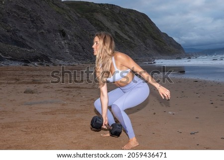 blonde woman doing squats with a dumbbell on the seashore at sunset