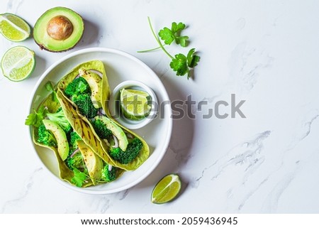 Green vegan tacos with broccoli, avocado and green flat bread in a white bowl, white marble background, top view, copy space. Vegetarian fast food.