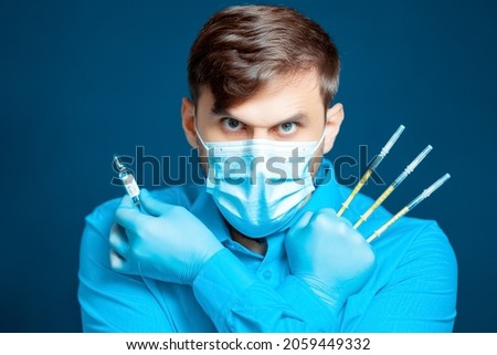 A doctor in a medical mask and gloves, in a blue uniform, poses with thin syringes between his fingers at the camera