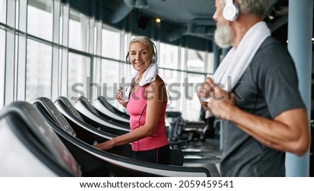 Side view elder smiling woman working out on treadmill, looking at camera, selective focus. Senior man in gray t-shirt, blured. Elder couple enjoying the moment. Modern gym on backgrund. Royalty-Free Stock Photo #2059459541