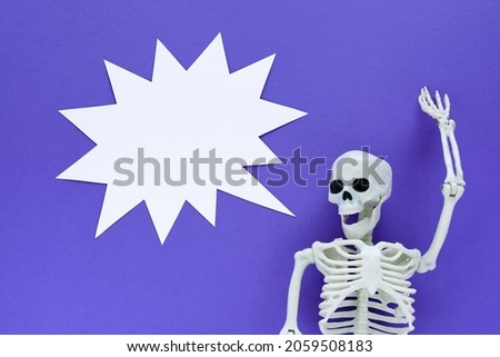 Skeleton on violet background with white blank paper thorny sound bubble. Anatomical plastic model human skeleton with one raised hand screams loudly. Empty yell cloud, copy space. Purple Halloween.