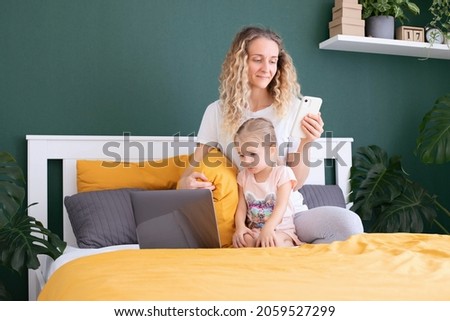 A young mother is sitting on the bed with a little girl with a phone in her hands, texting, sitting on social networks, shopping online, working online. Modern bedroom interior. Selective focus
