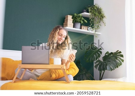 A young girl is lying, sitting on the bed with a phone in her hands, texting, sitting on social networks, shopping online, working online. Modern bedroom interior. Selective focus