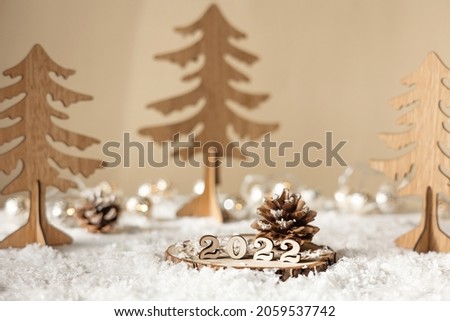 New year 2022. Numbers 2022 on wooden stand on beige pastel blurred background with decorative fir trees, snow and lights. Christmas greeting card.