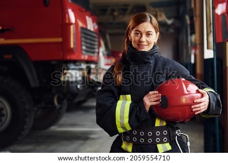 Holds red hat in hands. Female firefighter in protective uniform standing near truck.