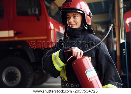 Holds extinguisher in hands. Female firefighter in protective uniform standing near truck.
