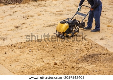 Worker in use vibratory plate compactor for path construction. Plate compactor for compaction sand. Industrial equipment with a combustion engine. Royalty-Free Stock Photo #2059562405