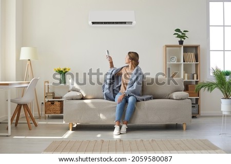 Woman who's sitting on sofa under warm plaid in living room switches off her air conditioner on wall. Young girl adjusting modern AC system, regulating temperature and enjoying cool fresh air at home Royalty-Free Stock Photo #2059580087