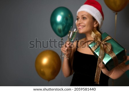 Focus on Christmas present in green shiny wrapping gift paper and gold bow in the hand of a smiling woman in Santa Claus hat drinking champagne and celebrating new year party with balloons background