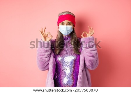 Covid-19, social distancing and fashion concept. Fashionable asian senior woman in face mask and glitter dress, showing okay signs in approval, pink background
