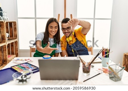 Young hispanic couple using laptop sitting on the table at art studio smiling making frame with hands and fingers with happy face. creativity and photography concept. 