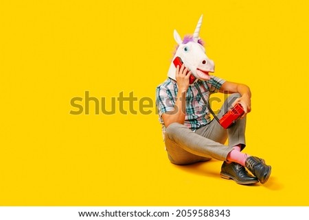 a young man wearing a unicorn mask talking on the phone, using a colorful red landline telephone, sitting on a yellow background Royalty-Free Stock Photo #2059588343