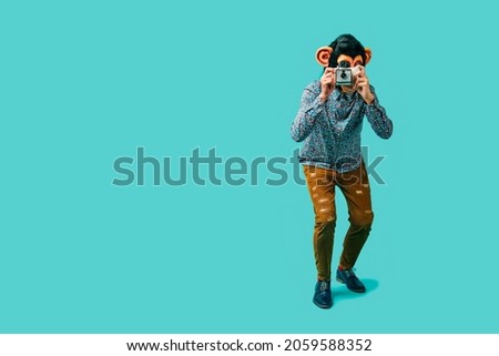 a young man, wearing a monkey mask, takes a picture with a retro instant camera, standing on a blue background with some blank space on the left