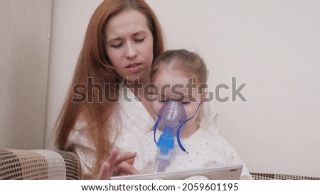 Mom treats her daughter with inhalation together they watch cartoons on tablet. Family treating child at home for colds. Child suffers from cough. Kid is receiving respiratory therapy with nebulizer.