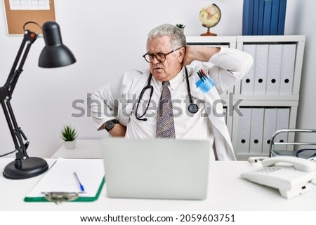 Senior caucasian man wearing doctor uniform and stethoscope at the clinic suffering of neck ache injury, touching neck with hand, muscular pain 