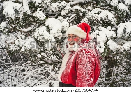 Happy senior man in traditional red Santa Claus costume standing near snow-covered tree branches, looking at the camera and laughing.