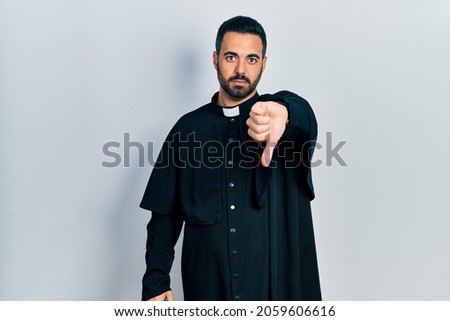 Handsome hispanic man with beard wearing catholic priest robe looking unhappy and angry showing rejection and negative with thumbs down gesture. bad expression. 