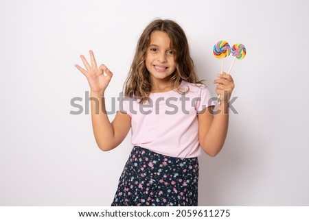 Beautiful little girl with lollipop and okay sign isolated over white background.