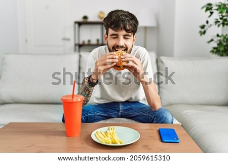 Young hispanic man eating classical burger and drinking soda sitting on the sofa at home.