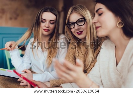 Group of smiling female friends in casual clothes sitting at wooden table and browsing smartphone while working together on project