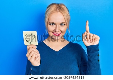 Young blonde woman holding heart reminder smiling with an idea or question pointing finger with happy face, number one 