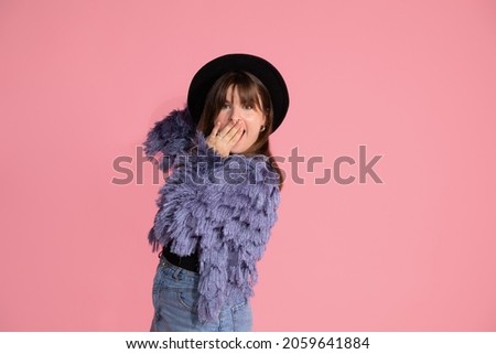  young, beautiful dark-haired woman in black hat, in surprise, covered her mouth with hand. On pink background in studio. 