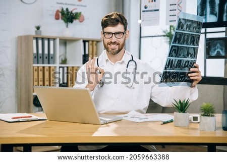 Yes, I did it. Likable smiling male doctor scientist, showing okay sign on camera while sitting at desk with x ray scan in hands. Bearded doctor using laptop at office.