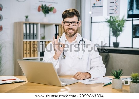 Caucasian male doctor showing approval okay gesture and smiling on camera while sitting at modern office with laptop on table. Modern gadget for medical workers Positive human emotions.