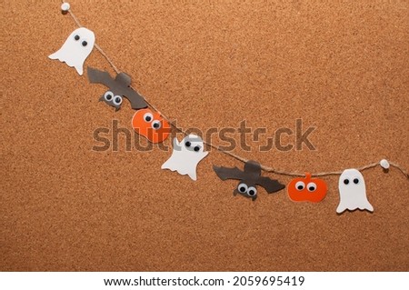Homemade paper garland for Halloween in the form of pumpkins, ghosts and bats with eyes on a jute rope attached with buttons to a cork board with a place for text