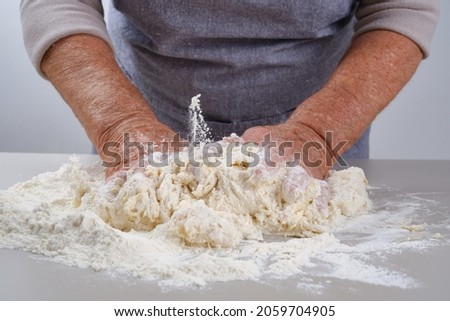 Female hands of an elderly woman knead the dough on a gray table.  Senior woman in a gray apron preparation of dough for a pizza, bread, pasta, festive cake. Shallow depth of field