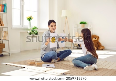 Professional children's psychologist talking to little kid about emotions. Girl chooses happy smiley emoticon from two EQ cards that female therapist shows her during interview appointment meeting