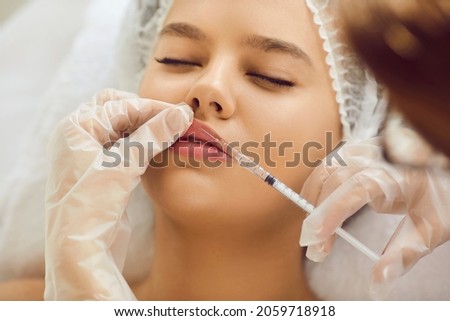 Beautiful woman getting professional treatment at clinic of aesthetic medicine. Young lady receiving botox injection for bigger, plumper lips. Doctor's hands holding syringe above face in close up Royalty-Free Stock Photo #2059718918