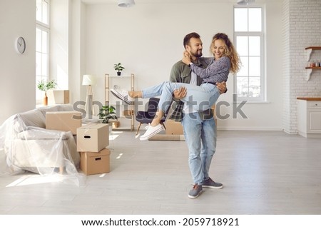 Happy young married couple on threshold of new life. Newlyweds having fun in big new home. Strong man carrying his woman in arms in newly bought modern spacious unfurnished house. Real estate concept Royalty-Free Stock Photo #2059718921