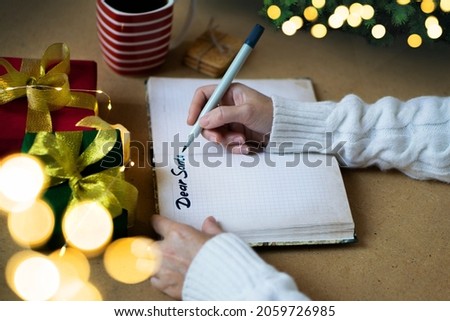 Woman's hands in white sweater writing letter to Santa in notebook, selective focus, bokeh lights, atmospheric photo. Christmas shopping and gifts concept.