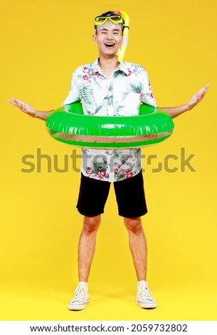 Young attractive Asian man wearing hat and white Hawaiian shirt wearing yellow snorkel mask and green swim ring around his waist against yellow background. Concept for beach vacation holiday.