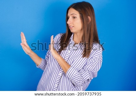 Displeased Young european brunette woman wearing stripped shirt on blue background keeps hands towards empty space and asks not come closer sees something unpleasant