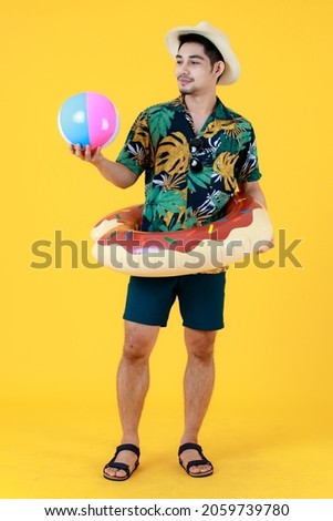 Smiling young Asian man in printed Hawaiian shirt and swim ring on waist looks at a colorful beach ball. Full body studio portrait on yellow background. Summer holiday travel preparation concept