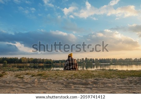  Girl traveler in autumn sits in a plaid blanket on the shore and looks at the sunset