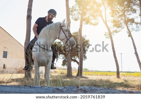 A rider, a Caucasian man with a beard, is riding his white horse, which is eating herbs, in front of an old house and next to the trunks of some trees in the middle of a field.