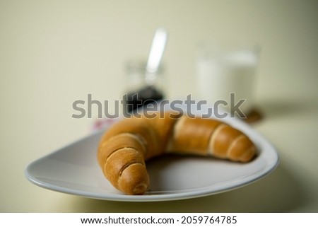 Twisted baked croissant bagel with a cup of milk and a jar of jam on a plate and checkered cloth with white background