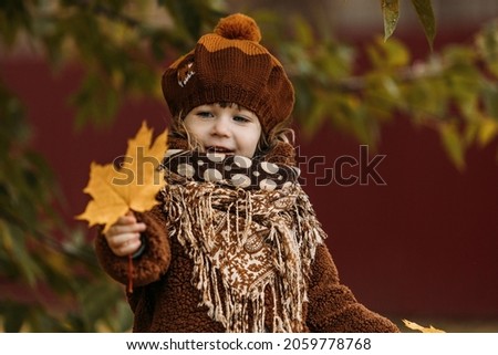 A little girl, a child in a brown fur coat, and in a brown beret walks in autumn with yellow maple leaves.