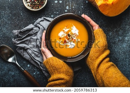 Female hands in yellow knitted sweater holding a bowl with pumpkin cream soup on dark stone background with spoon decorated with cut fresh pumpkin, top view. Autumn cozy dinner concept  Royalty-Free Stock Photo #2059786313
