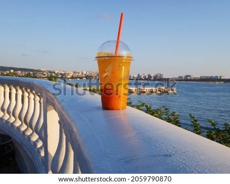a transparent plastic glass with a cocktail stands on a white fence on the embankment against the background of the sea and the evening sky. 