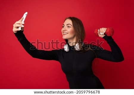 Photo of beautiful smiling positive adult brunette woman wearing black sport clothes white headphones nad holding red dumbbell standing isolated over red background using mobile phone taking selfie