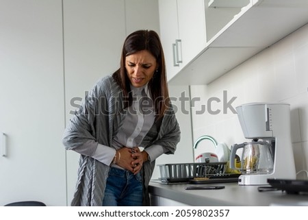 Unhappy sick woman with hands holding pressing her crotch lower abdomen. Medical or gynecological problems, healthcare concept. Mid adult woman suffering from abdominal pain while standing in kitchen. Royalty-Free Stock Photo #2059802357