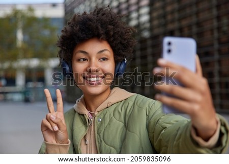 Smiling good looking young woman smiles broadly takes self portrait makes selfie on good quality smartphone camera listens favorite music in hedphones dressed in hoodie and jacket poses in downtown