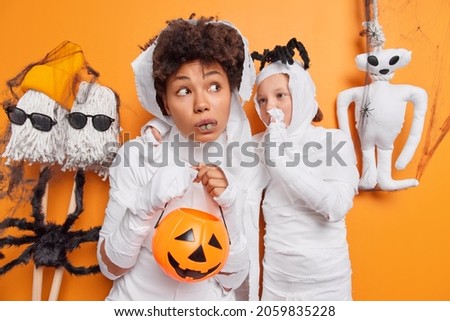 Small child whispers secret to her mum wear carnival costumes for halloween celebration hold carved pumpkin surrounded by spooky creatures against orange background. 31st of October is coming